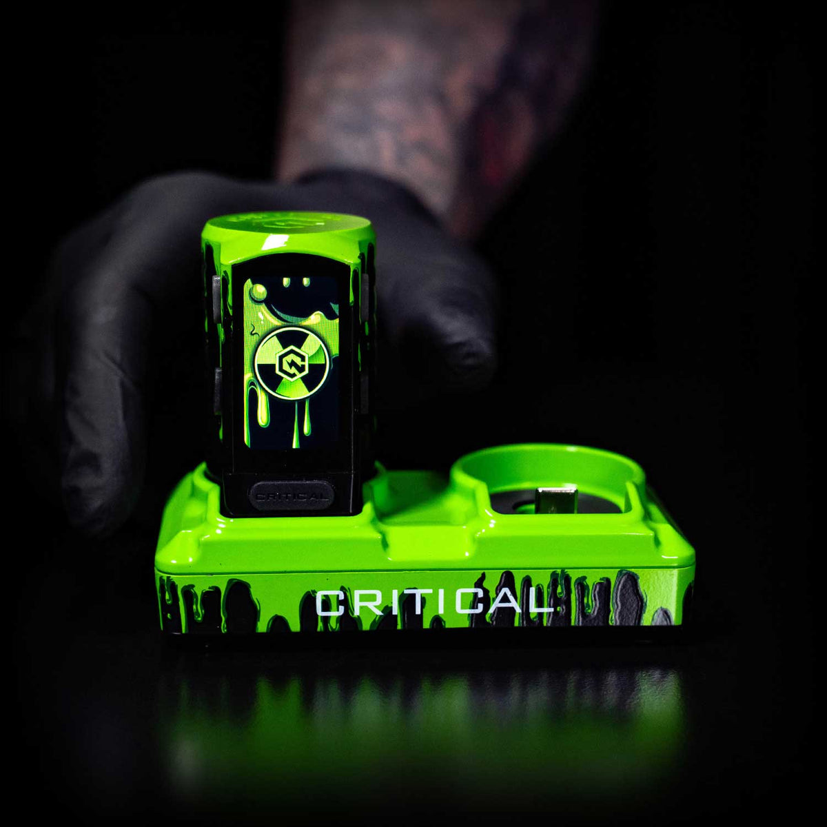 Green tattoo machine with 'Critical' branding held by a person wearing black gloves against a dark background.